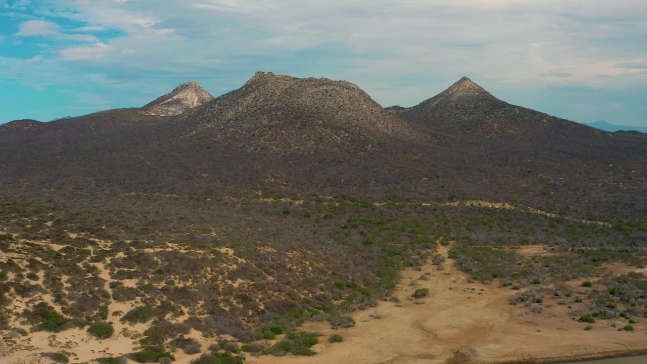 Mid-clip aerial view in Laguna Beach, Baja California Sur, showing sandy foreground terrain, patchy shrubs, a dark mountain ridge, rocky peaks, blue sky, and no visible people.