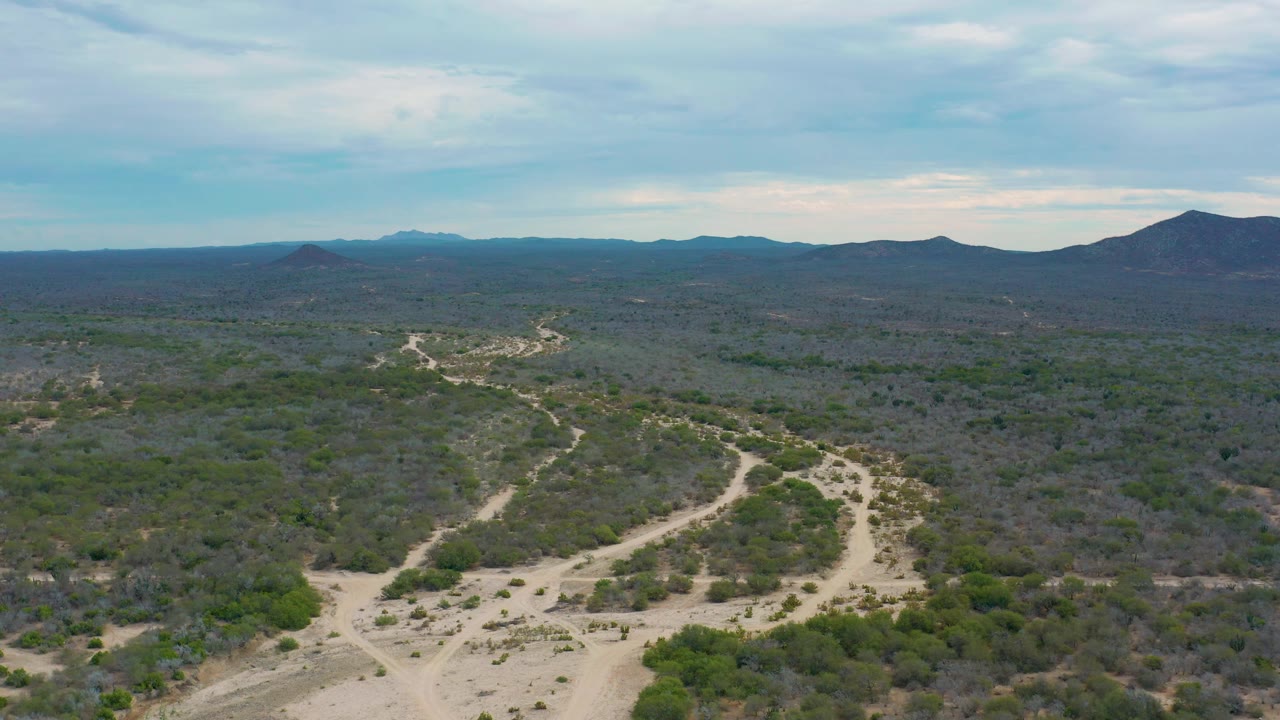 Mid-clip aerial view over Laguna Beach, Baja California Sur, Mexico, where sandy tracks cut through green and gray brush, with a broad mountain horizon beneath muted blue cloud cover, no visible people.