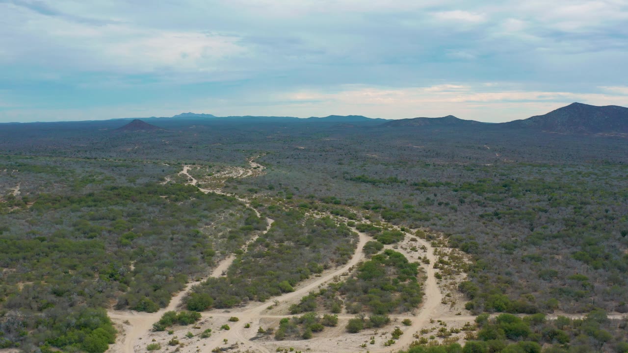 Mid-clip aerial frame from Laguna Beach, Baja California Sur, with the winding dirt road set lower in frame, wide scrubland beyond it, and no visible people.