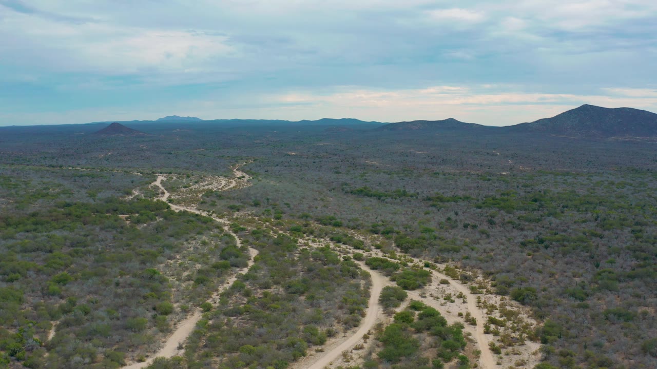 Aerial view in Laguna Beach, Baja California Sur with the sandy wash curving from the lower frame through sparse scrub toward a broad dark hill on the right, with no visible people.