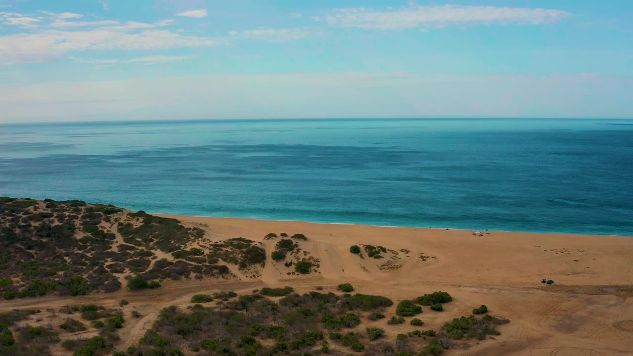 Laguna Beach shoreline in Baja California Sur appears from above, with turquoise water shifting darker offshore, tan sand below, scrub vegetation at the lower edge, and no visible people.