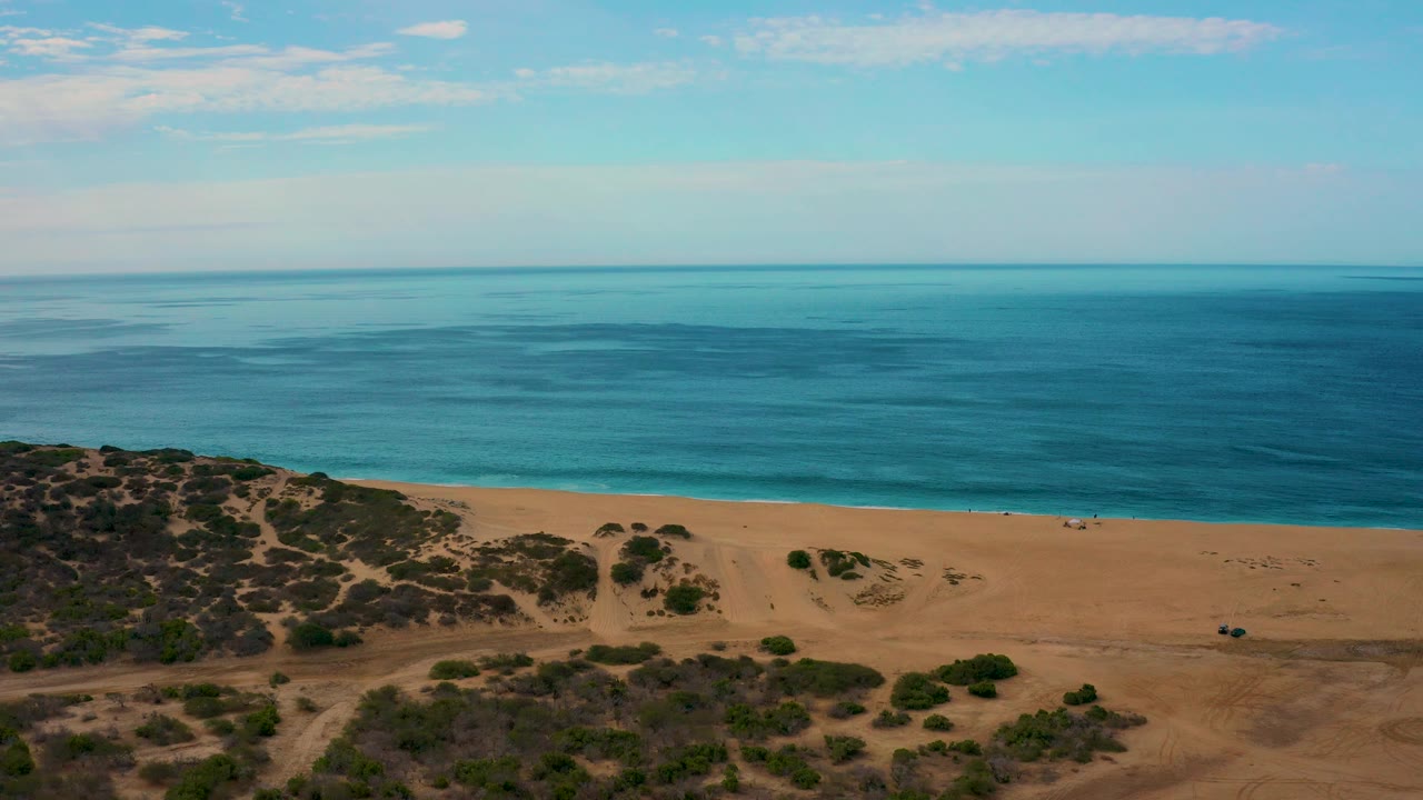 A high aerial frame over Laguna Beach, Baja California Sur shows a level ocean horizon, narrow surf line, broad tan sand, sparse vegetation, and no visible people.