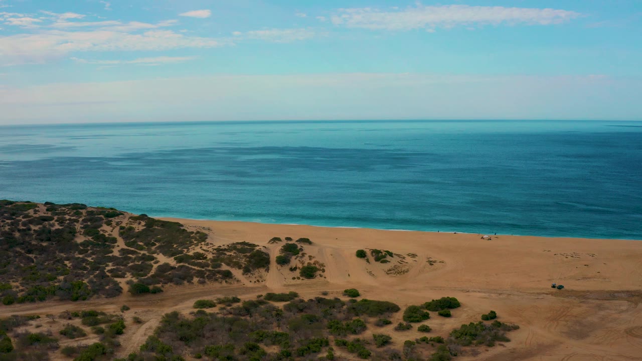 Mid-clip frame of Laguna Beach in Baja California Sur with calm blue water across the upper frame, a wide band of sand, scrubland in front, light cloud cover, and no visible people.