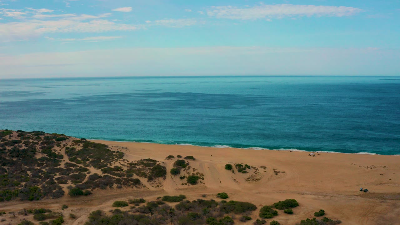 Aerial frame over Laguna Beach, Baja California Sur, with blue ocean beyond a tan shoreline, scrub dunes at lower left, and no visible people.