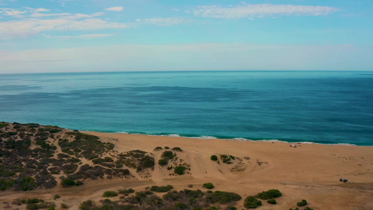 Wide aerial frame of Laguna Beach, Baja California Sur, with calm blue sea, empty tan sand, low vegetation near the bottom edge, and no visible people.