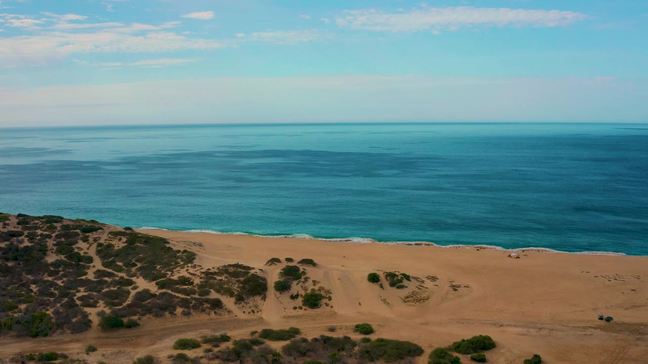 Aerial frame over Laguna Beach, Baja California Sur, with the ocean horizon high in frame, a sandy beach below, scrubby dune slope at lower left, and no visible people.