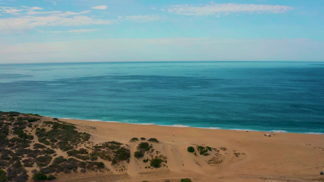 The second frame keeps the Laguna Beach coastline in a high drone-style composition, with dune paths below, tan sand across the middle, turquoise water near shore, and no visible people.