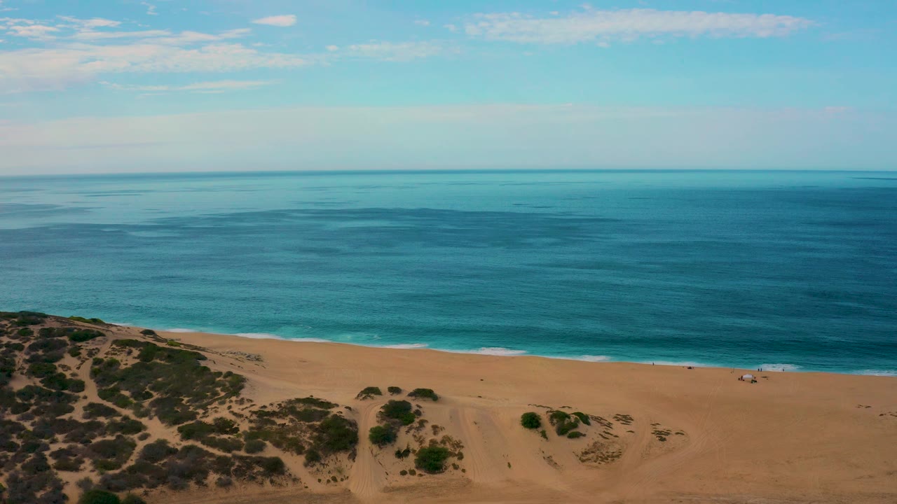 Aerial shoreline detail at Laguna Beach shows darker scrub patches in the lower left, open tan sand, a thin broken surf line, blue ocean, and no visible people.
