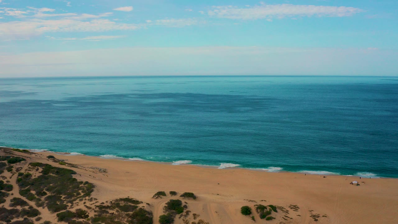 Opening frame of Laguna Beach, Baja California Sur, with blue ocean beyond a tan sandy shoreline, scrubby dunes at lower left, thin clouds overhead, and no visible people.