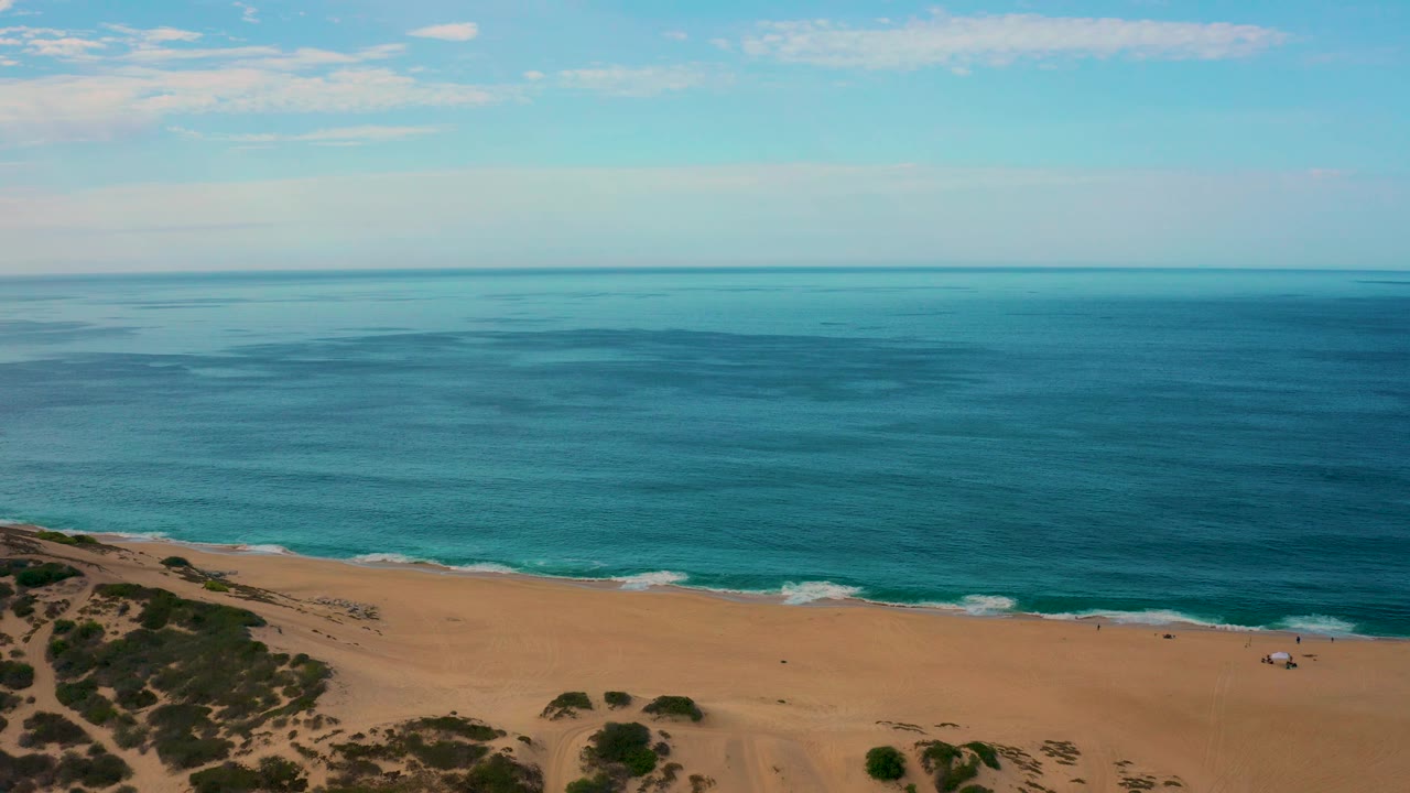 Aerial coastal view in Laguna Beach, Baja California Sur, showing a long tan beach under blue water, small white waves near shore, dune vegetation at left, and no visible people.