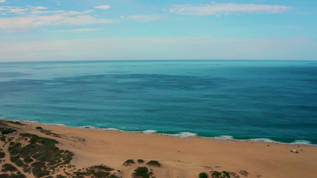 Laguna Beach shore seen from above with a wide expanse of blue ocean, a narrow line of foam along the beach, scrub-covered sand at lower left, and no visible people.