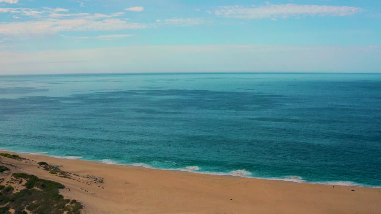 Opening aerial frame over Laguna Beach, Baja California Sur, with blue ocean, sandy shoreline, dune vegetation at lower left, thin clouds, and no visible people.