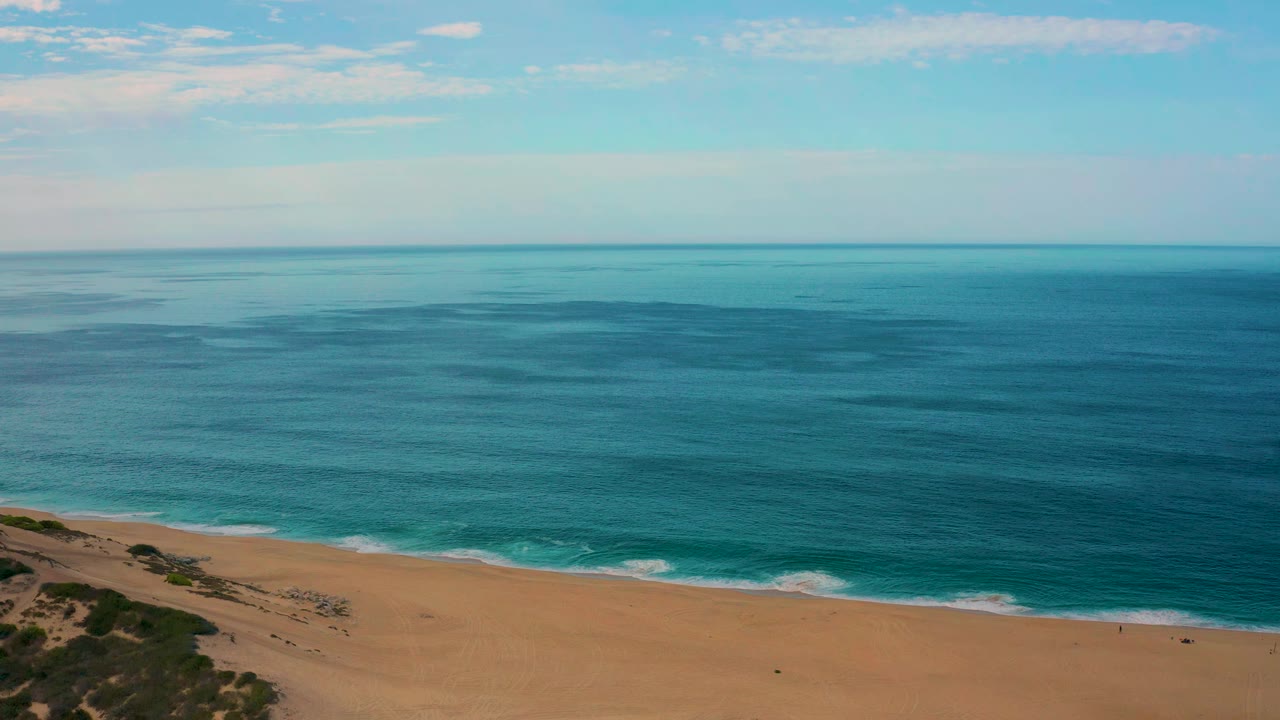 Aerial view one second in over Laguna Beach, Baja California Sur, where the shoreline sits lower in frame and the calm blue water stretches to a level horizon with no visible people.
