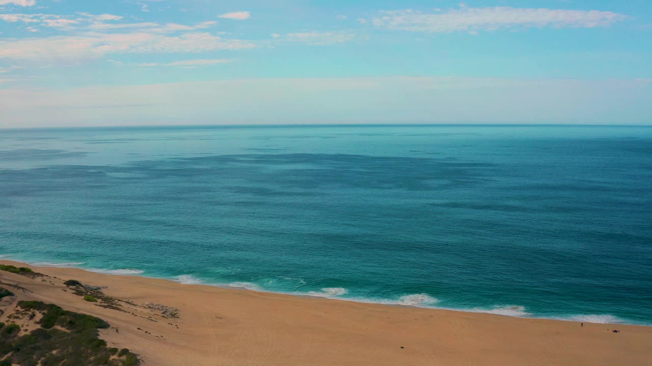 Aerial frame over Laguna Beach, Baja California Sur, with turquoise water meeting tan sand, small foam lines along the shore, open sky above, and no visible people.
