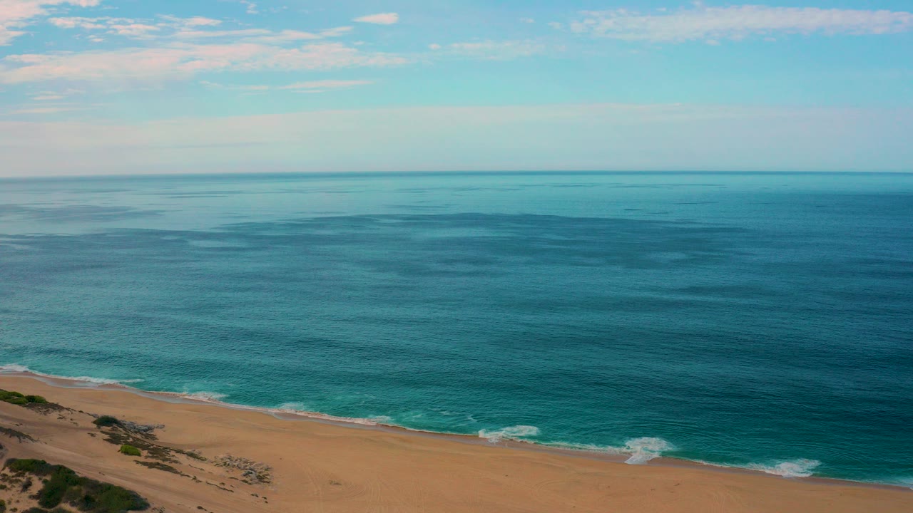 Aerial view over Laguna Beach, Baja California Sur, with blue ocean filling most of the frame, tan sand at the lower left, thin clouds above the horizon, and no visible people.