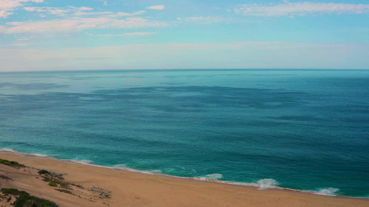 Elevated view of Laguna Beach, Baja California Sur, with darker blue ocean patches offshore, gentle white waves reaching the sand, pale sky overhead, and no visible people.
