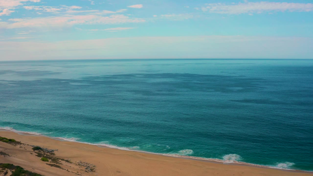 Aerial frame from Laguna Beach, Baja California Sur, with the beach crossing the lower left, turquoise water near shore, deeper blue water beyond, open sky, and no visible people.
