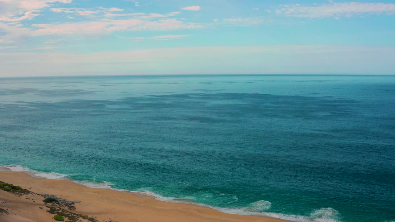 Aerial frame over Laguna Beach, Baja California Sur, with turquoise water, a sandy shoreline along the lower edge, pale clouds above the horizon, and no visible people.