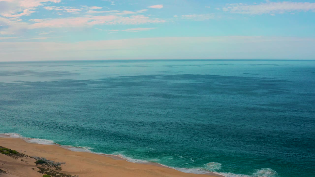 Aerial frame from Laguna Beach, Baja California Sur, with darker blue water patches, a sandy coast at the lower left and lower edge, a clear horizon, and no visible people.