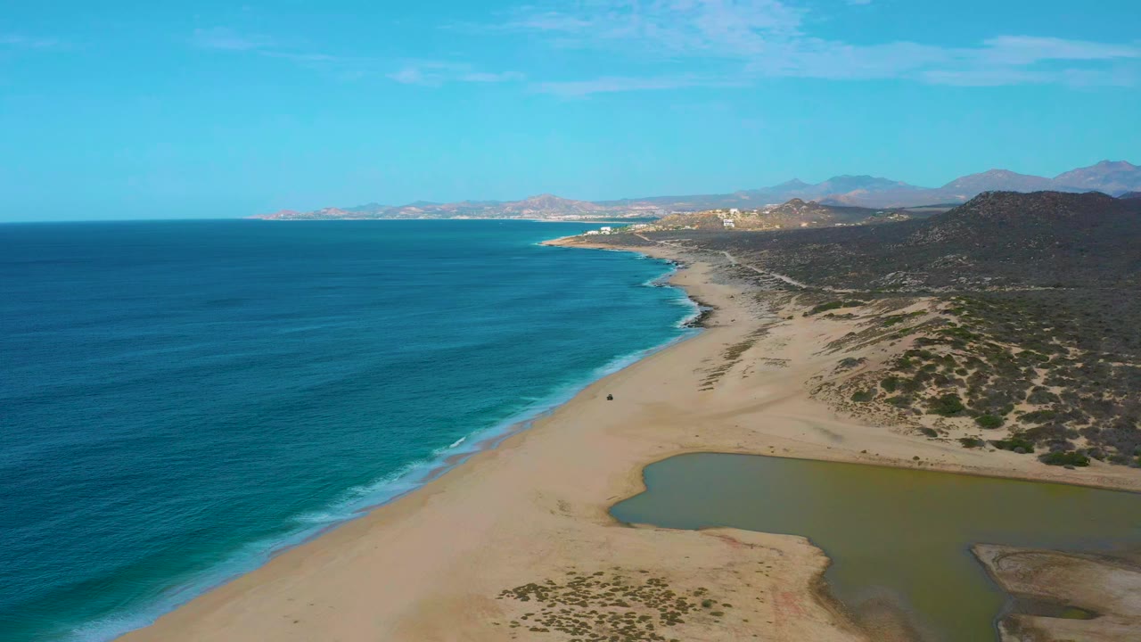 Aerial view over Laguna Beach, Baja California Sur, with blue ocean on the left, a broad sandy beach, a green lagoon, coastal scrub, and distant hills under clear sky.