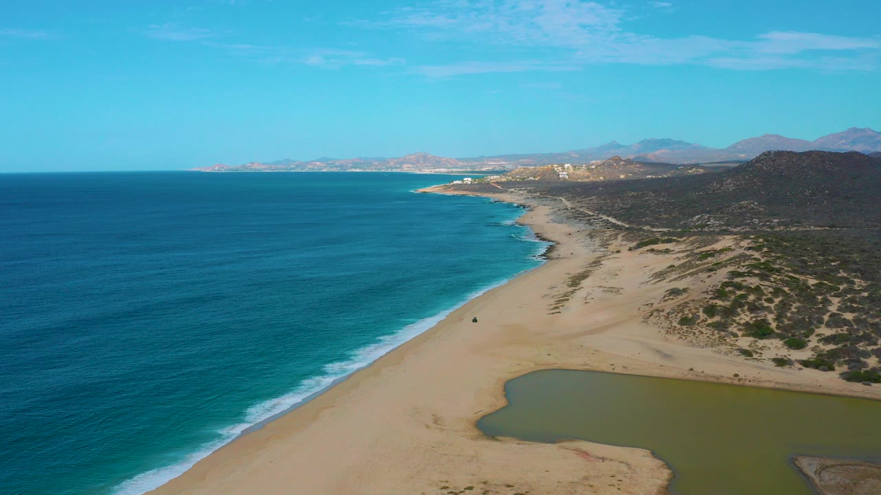 Opening aerial frame over Laguna Beach, Baja California Sur, with deep blue ocean on the left, pale sand and scrub on the right, a green lagoon at the lower edge, distant mountains, and no visible people.