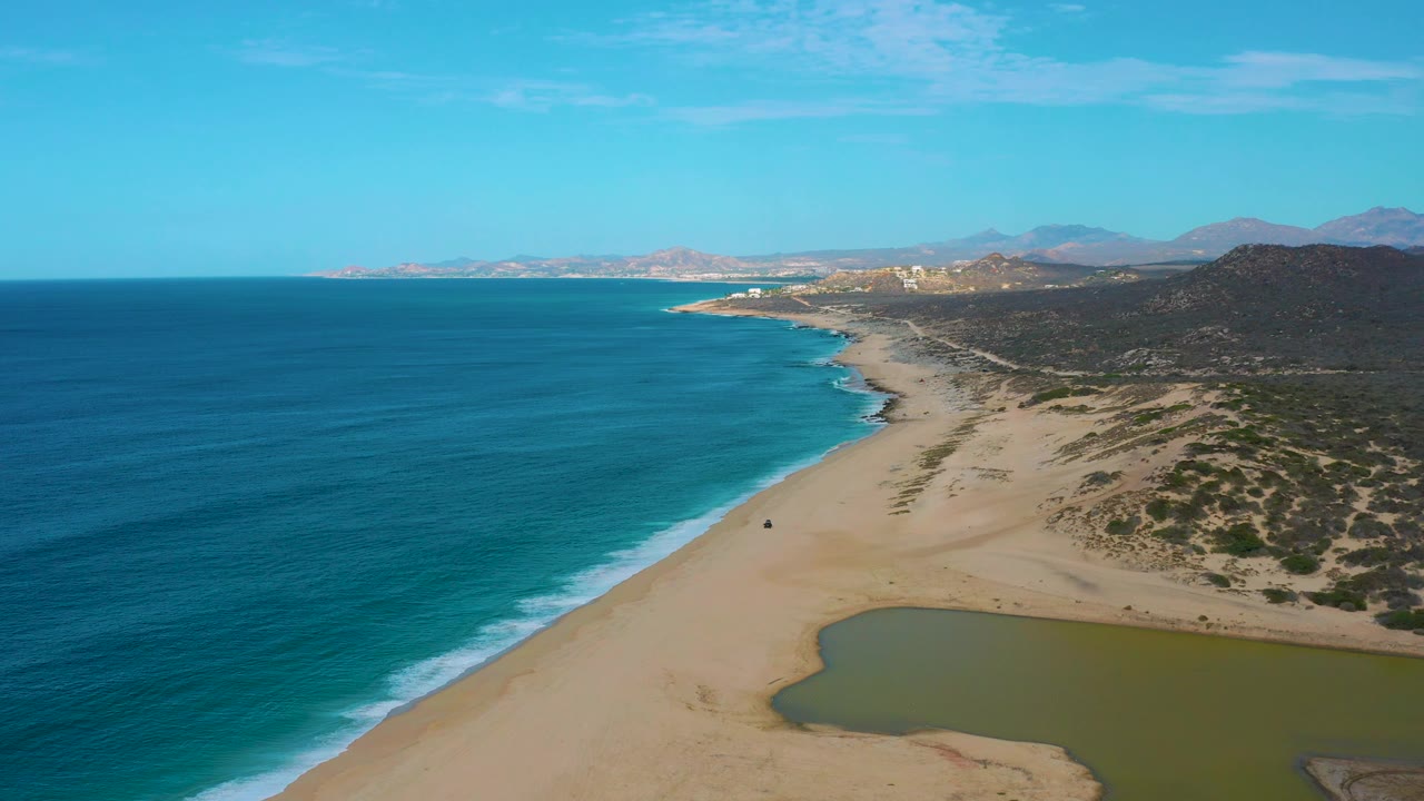 At 0:02, turquoise water and a narrow surf band run along the Laguna Beach sand while the lagoon edge sits lower right, backed by dark vegetation and hazy mountains, with no visible people.
