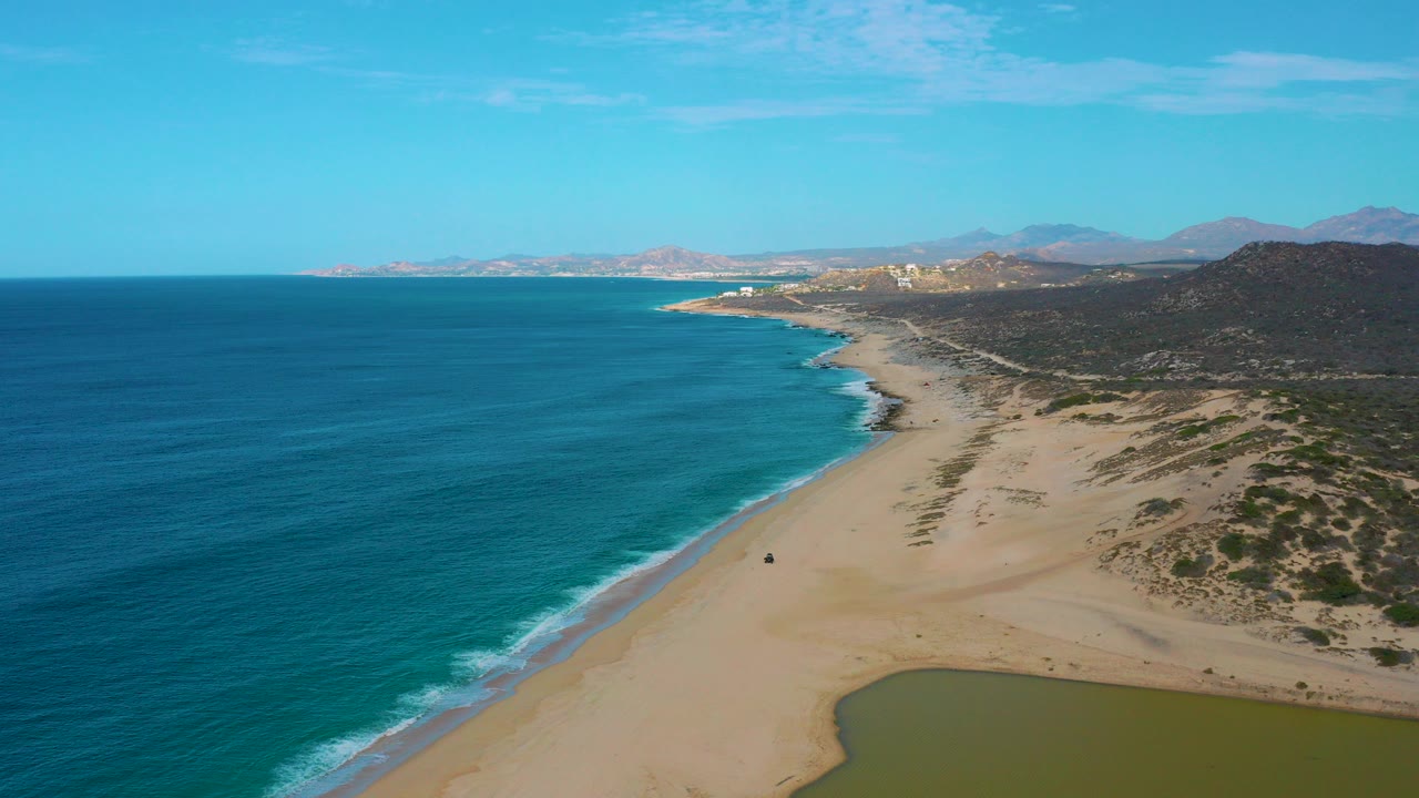 A high drone view over Laguna Beach, Baja California Sur shows turquoise ocean at left, a broad sandy shoreline at center, dunes and hills at right, distant mountains, and no visible people.