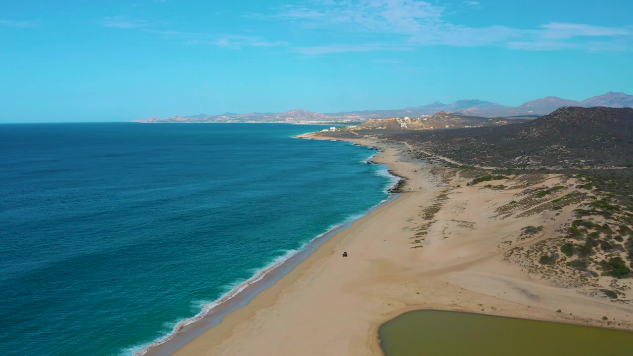 At 0:03, the Laguna Beach shoreline appears from above with calm blue water on the left, a broad empty beach across the center, scrub-covered dunes to the right, distant mountains, and no visible people.