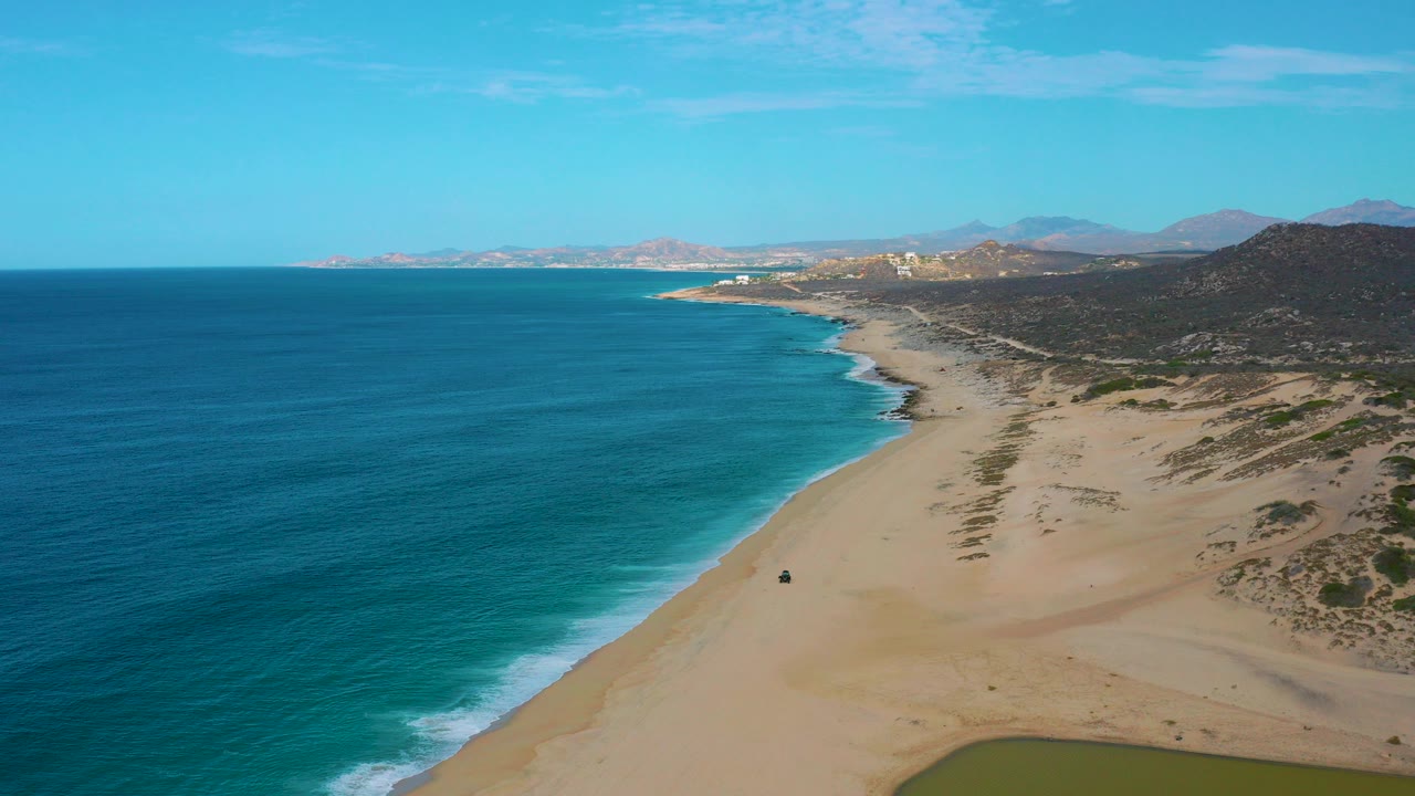 Opening aerial frame over Laguna Beach, Baja California Sur, with turquoise water on the left, a broad sandy shoreline below, distant hills on the right, and no visible people.