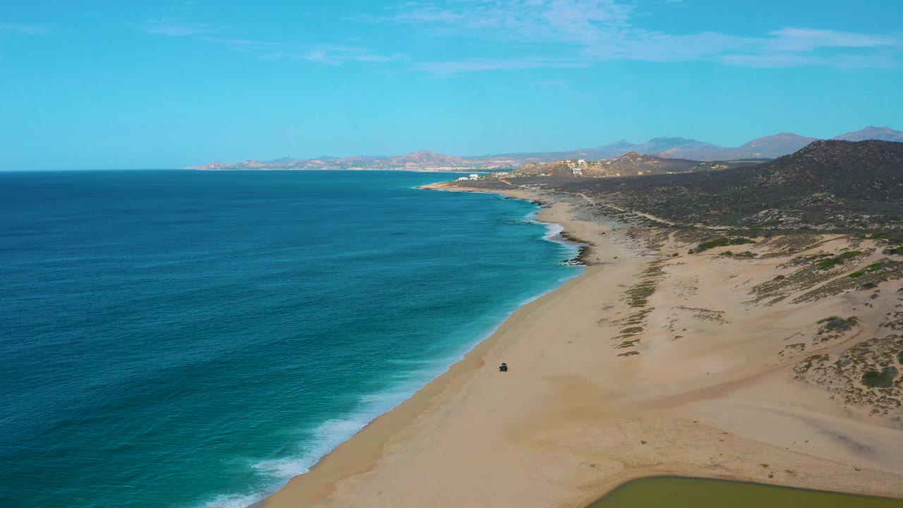 High oblique aerial frame of Laguna Beach, Baja California Sur, with the ocean filling the left half, empty sand and surf below, dark hills to the right, distant coast ahead, and no visible people.