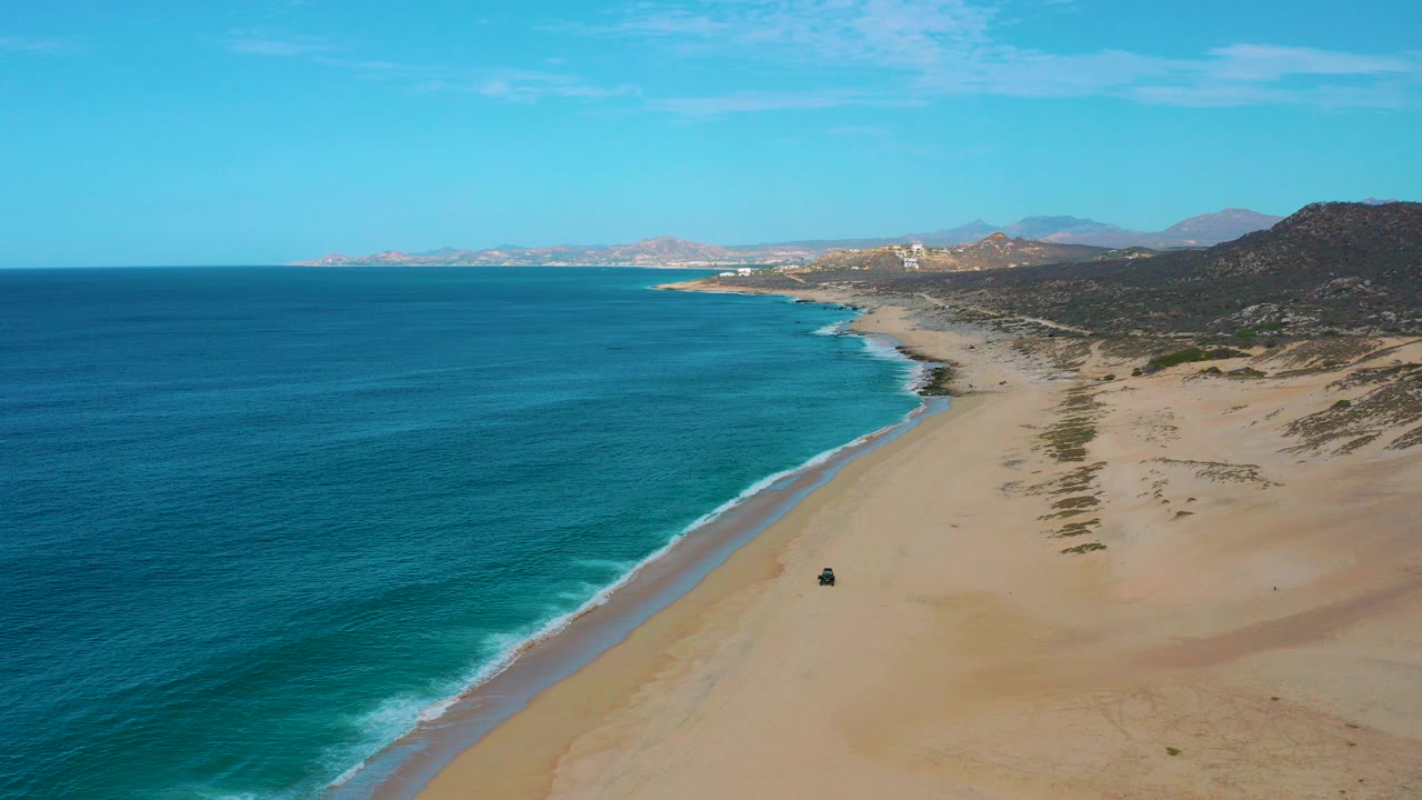 A high aerial frame over Laguna Beach, Baja California Sur shows deep blue ocean, pale sand, a thin surf line, distant coastline, inland hills, and no visible people.
