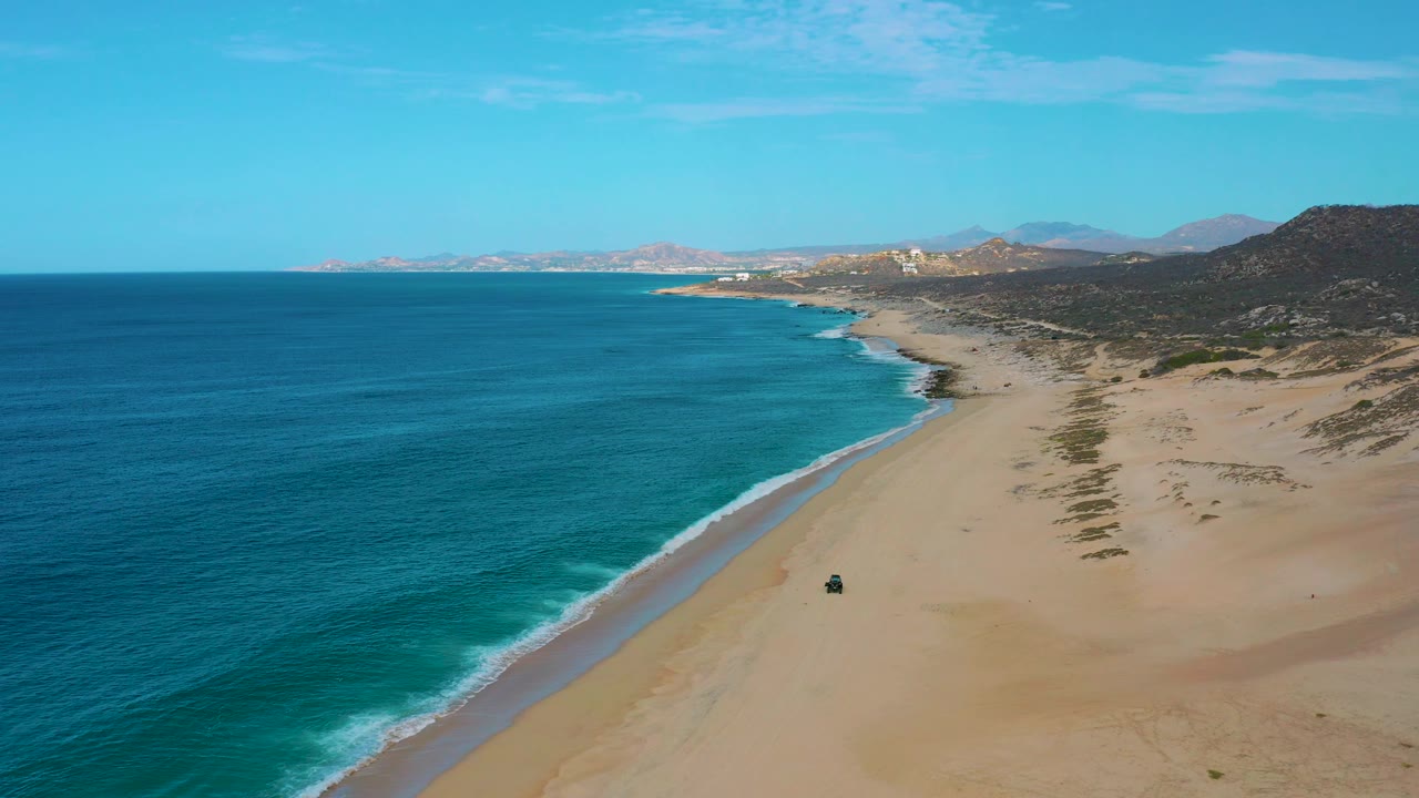 At 0:02 the frame shows the ocean taking most of the left side, the beach running diagonally into the distance, inland hills to the right, a small vehicle on the sand, and no visible people.