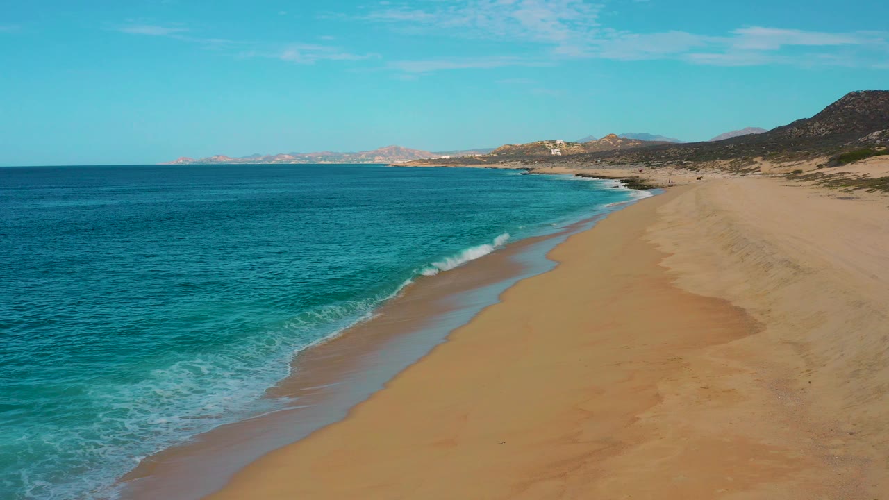 High drone frame above Laguna Beach, Baja California Sur, where blue-green ocean fills the left side and the tan beach curves toward distant rocky hills, with no visible people.