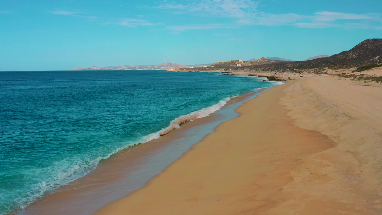 Drone frame over Laguna Beach, Baja California Sur, with a foamy wave breaking near the lower left, smooth empty sand to the right, and no visible people.