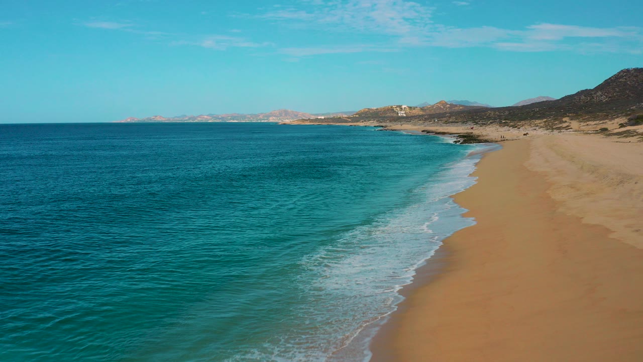 Aerial view over Laguna Beach in Baja California Sur, with turquoise ocean on the left, a band of white foam on the sand, distant hills on the horizon, and no visible people.