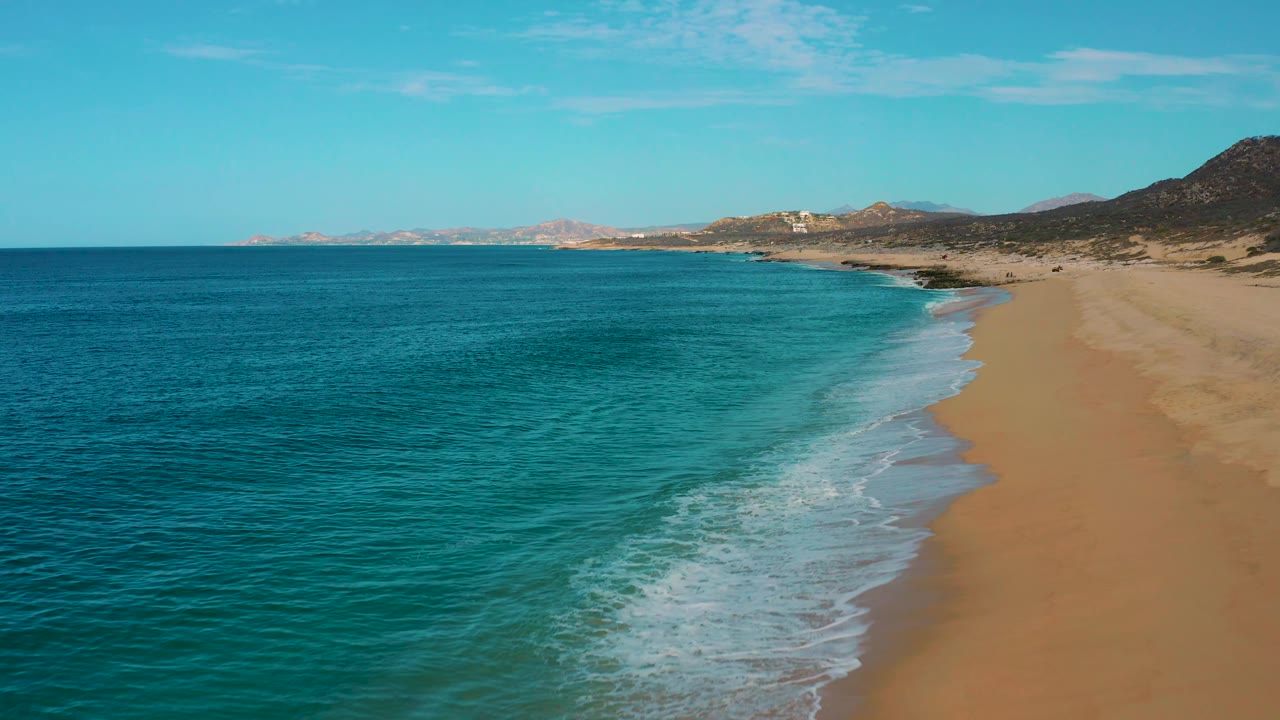 Aerial frame over Laguna Beach in Baja California Sur, with deeper blue ocean filling more of the foreground, a curved foamy wave line near the right, distant shoreline, and no visible people.
