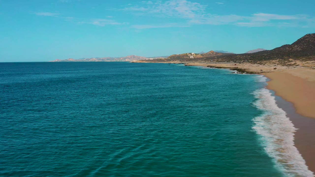 Aerial view in Laguna Beach, Baja California Sur, with blue-green sea filling the foreground, a curved surf line on the right, desert hills inland, and no visible people.
