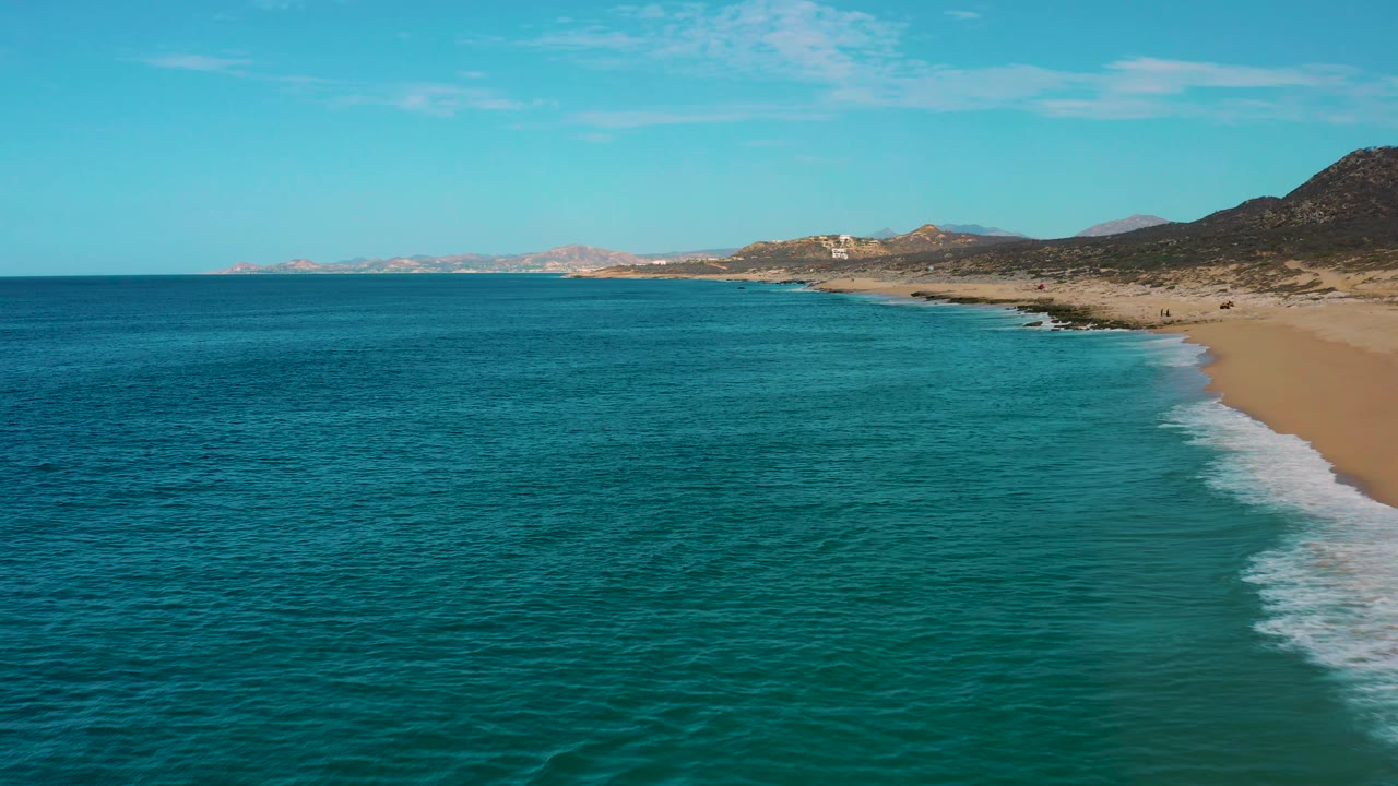 Laguna Beach aerial frame with textured turquoise ocean across most of the image, a narrow strip of surf and sand at right, hills in the background, and no visible people.