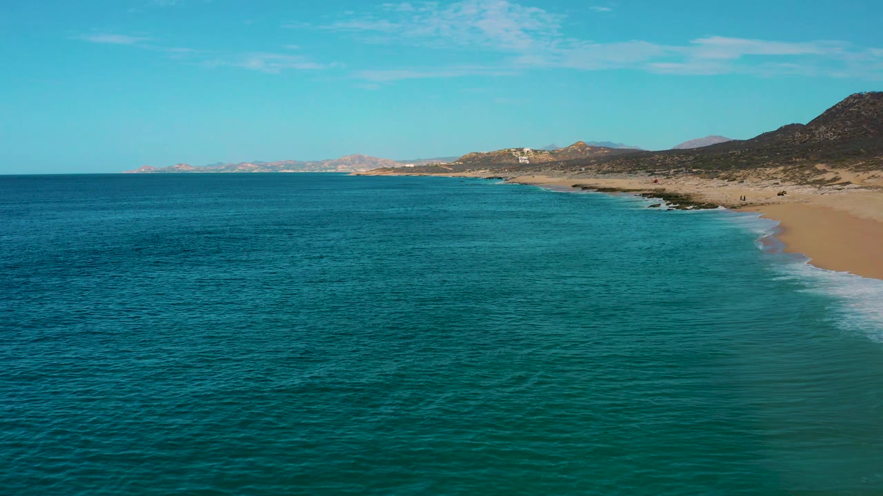 Mid-clip aerial over Laguna Beach, Baja California Sur, showing calm blue water, a pale surf band, open tan sand, low scrubby hills, and no visible people.