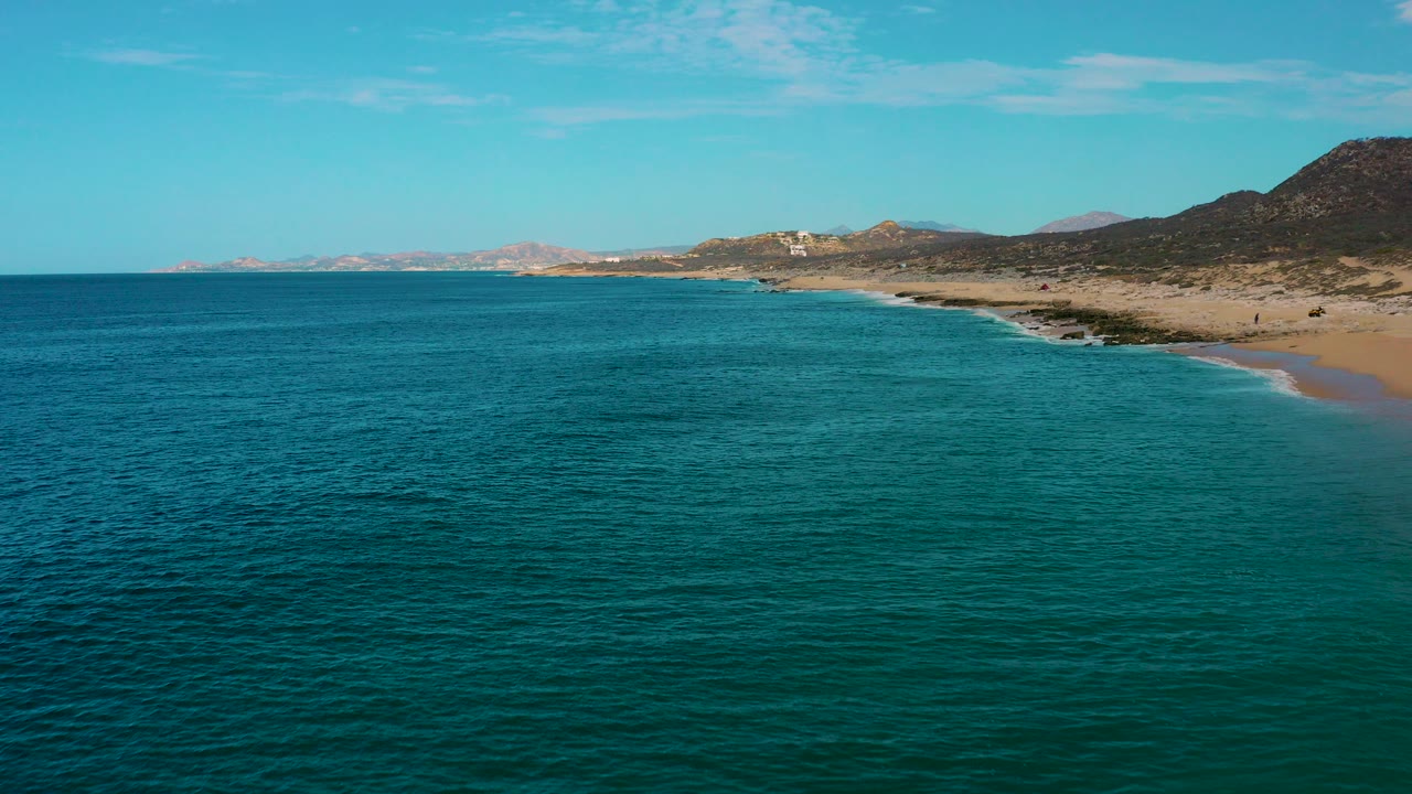 Aerial view over turquoise water toward Laguna Beach, Baja California Sur, with a sandy shoreline, low hills on the right, distant coast on the horizon, and no visible people.