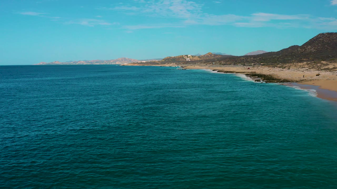 Aerial frame of Laguna Beach, Baja California Sur, showing textured teal ocean water, small waves near the sand, arid hills to the right, distant shoreline, and no visible people.