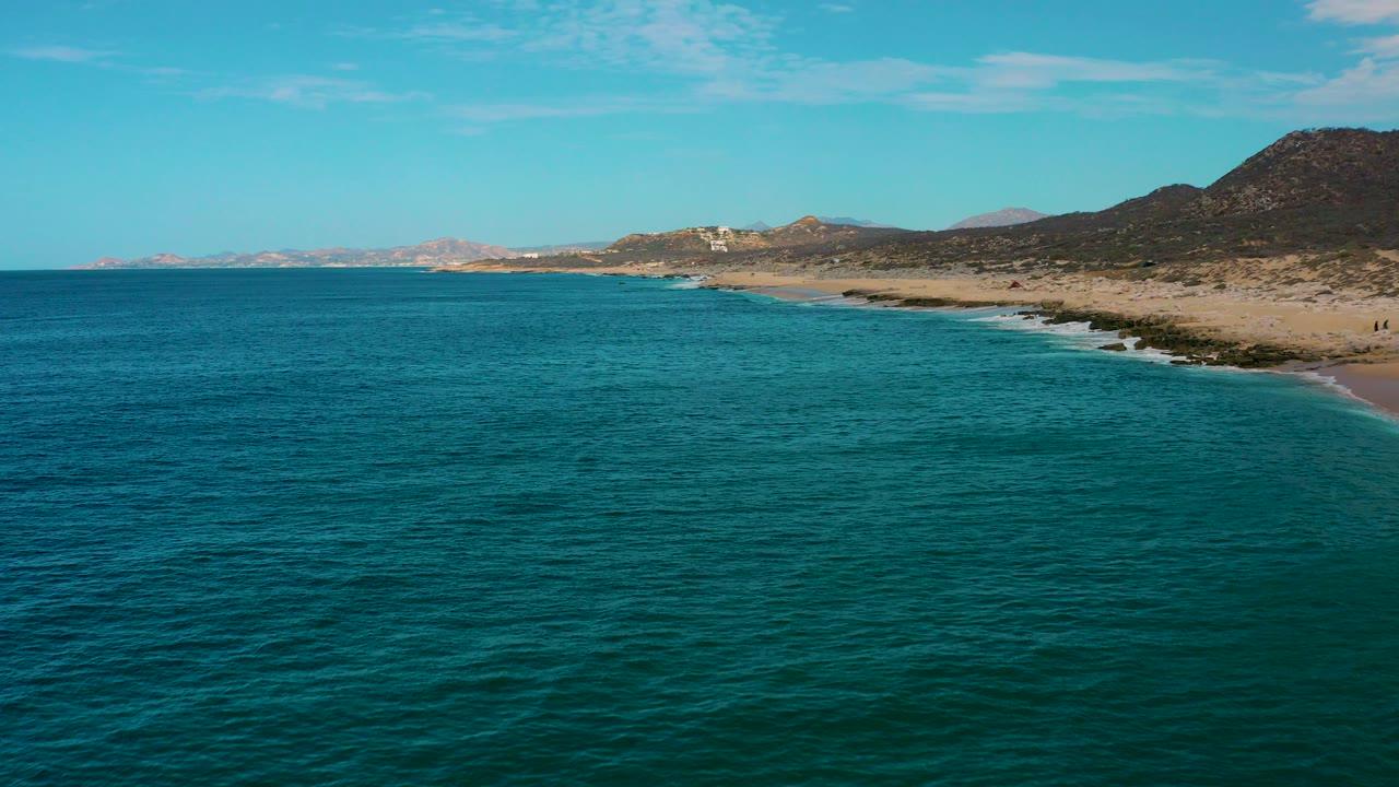 Laguna Beach, Baja California Sur coastline seen from above the blue green ocean, with sandy shore, low surf, arid hills, and no visible people.