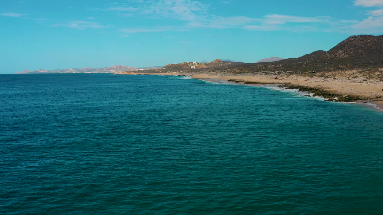 Blue green ocean fills most of the frame as the Laguna Beach shoreline curves along the right, with small white wave lines, dry hills, and no visible people.