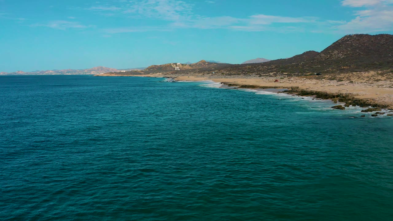 Aerial view at Laguna Beach, Baja California Sur, showing turquoise water across the foreground, a diagonal beach edge on the right, rocky surf lines, hills, and no visible people.