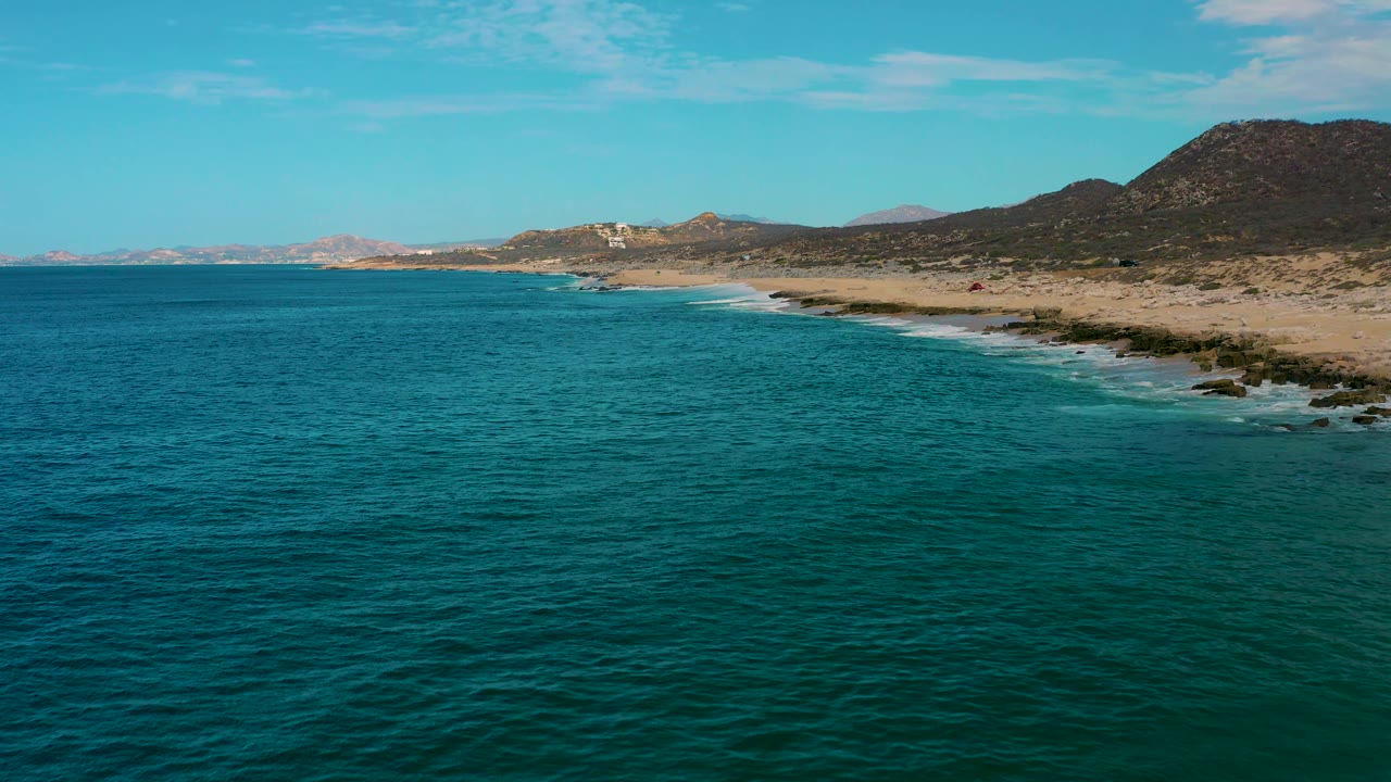 Wide aerial frame of Laguna Beach, Baja California Sur, with textured blue water in the foreground, pale sand and dark shoreline rocks at right, distant hills, and no visible people.