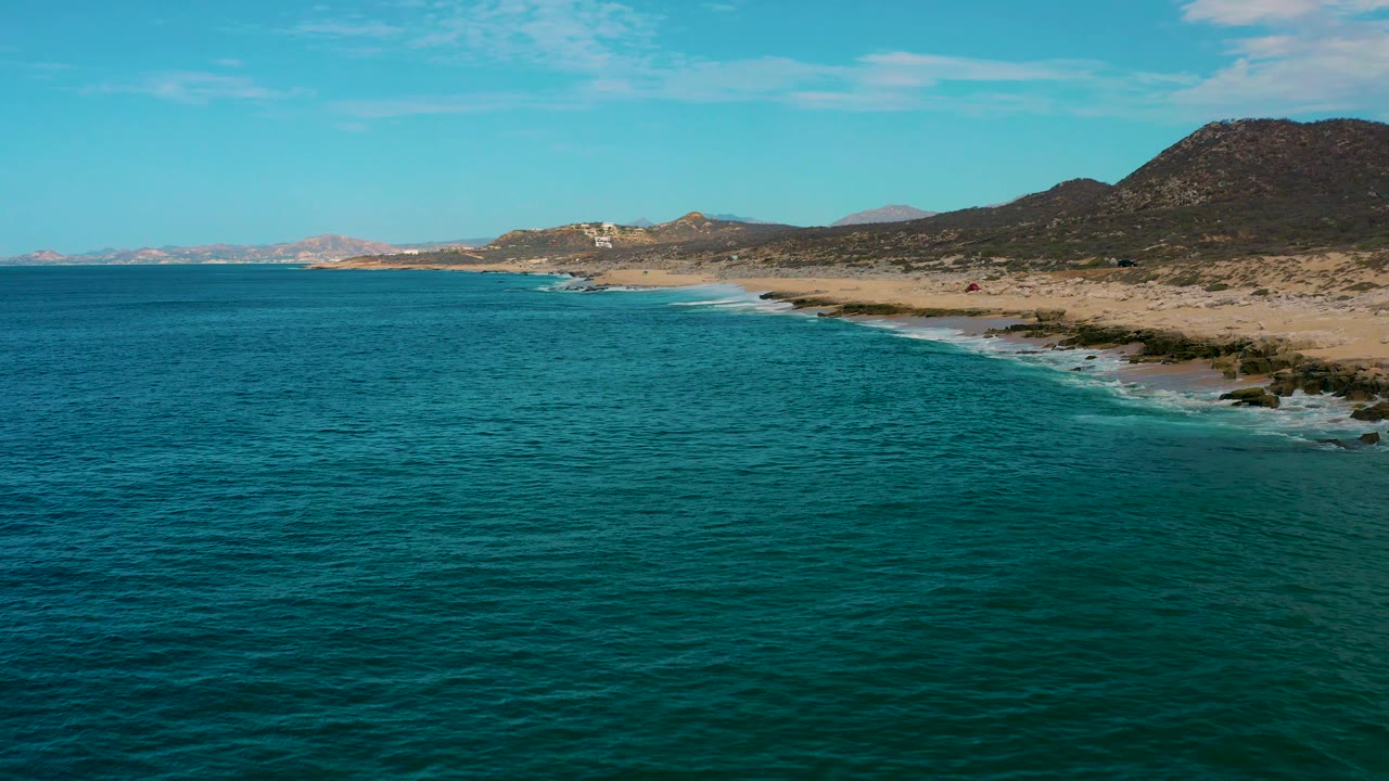 Aerial frame over Laguna Beach, Baja California Sur, where blue water fills most of the image and white wave lines trace the sandy, rocky shore below the hills, with no visible people.