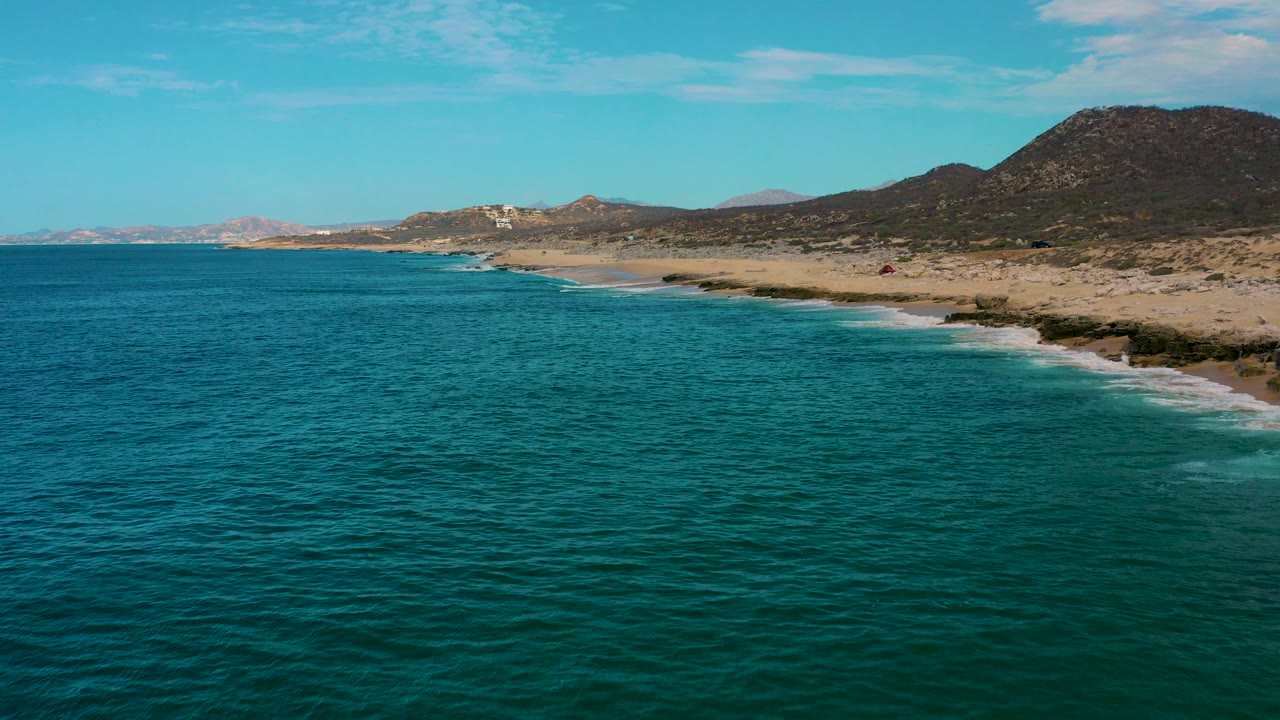 Aerial view of teal ocean beside a sandy coast and dark hills.