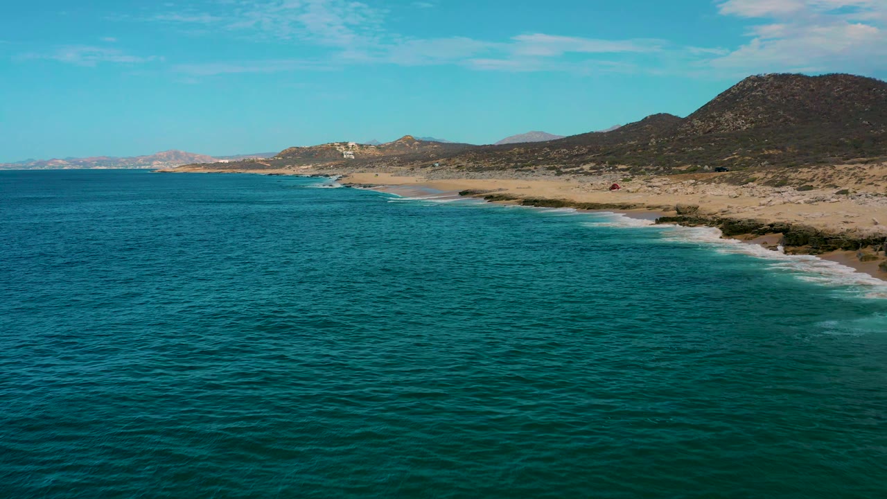 Laguna Beach, Baja California Sur frame showing teal water across the lower frame, a thin white surf line at the beach, low arid hills, and no visible people.