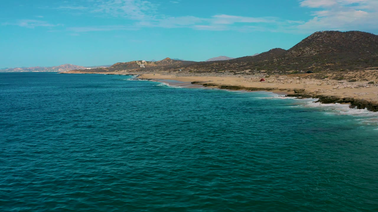 Laguna Beach, Baja California Sur frame with broad teal ocean foreground, a curving pale beach, white shore break, a large dark hillside at right, and no visible people.
