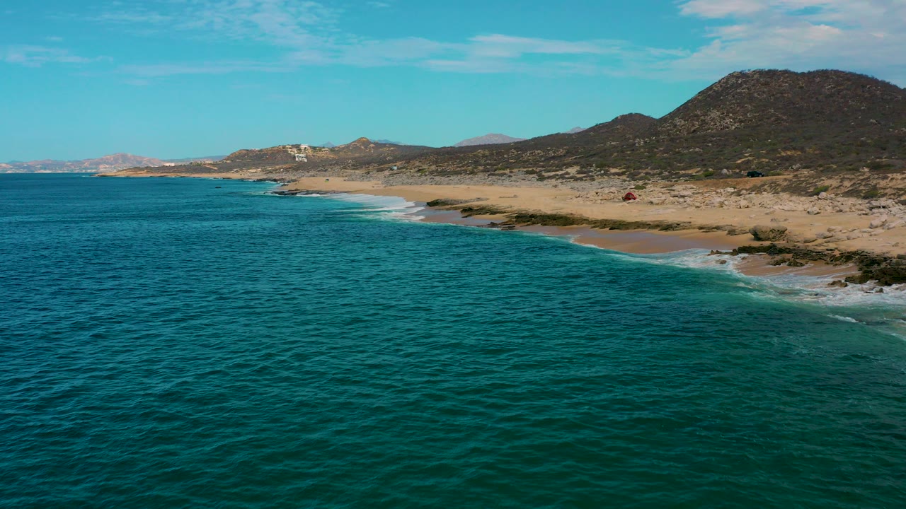 Opening aerial frame over Laguna Beach, Baja California Sur, with turquoise water in the foreground, a sandy shoreline to the right, rocky hills beyond, and no visible people.